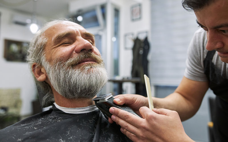 Barber trimming the beard of a relaxed senior man in a salon.