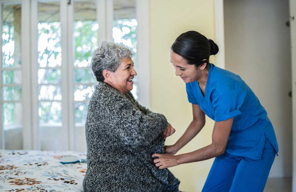 Caregiver in blue uniform helps elderly woman in gray sweater with daily grooming in a cozy room.