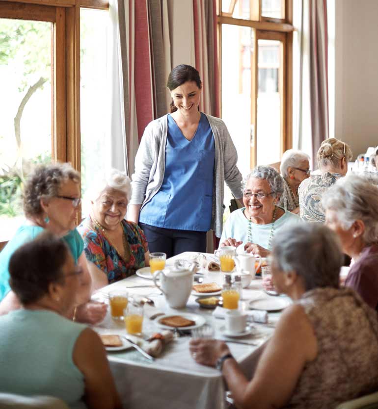 Caregiver with senior women smiling and dining together, enjoying breakfast at a communal table.