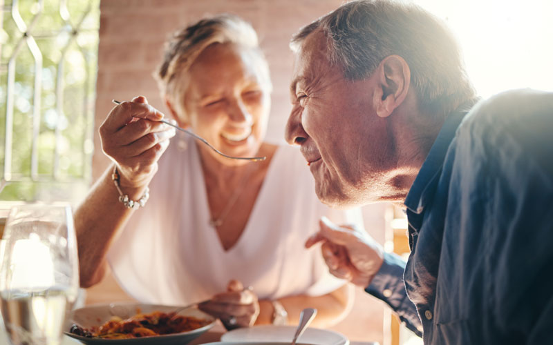Elderly woman feeding her smiling male companion in a cozy restaurant setting.