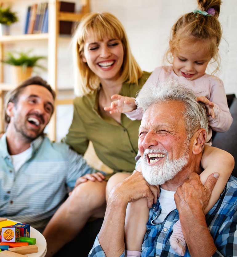 Elderly man joyfully playing with young girl, surrounded by family members who are laughing and smiling.