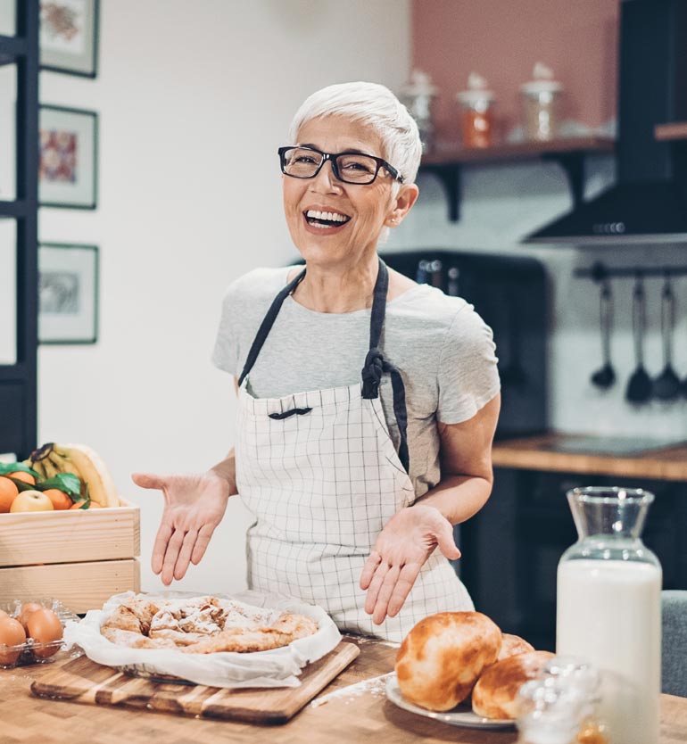 Elderly woman wearing glasses and apron smiling over freshly baked goods in a cozy kitchen.