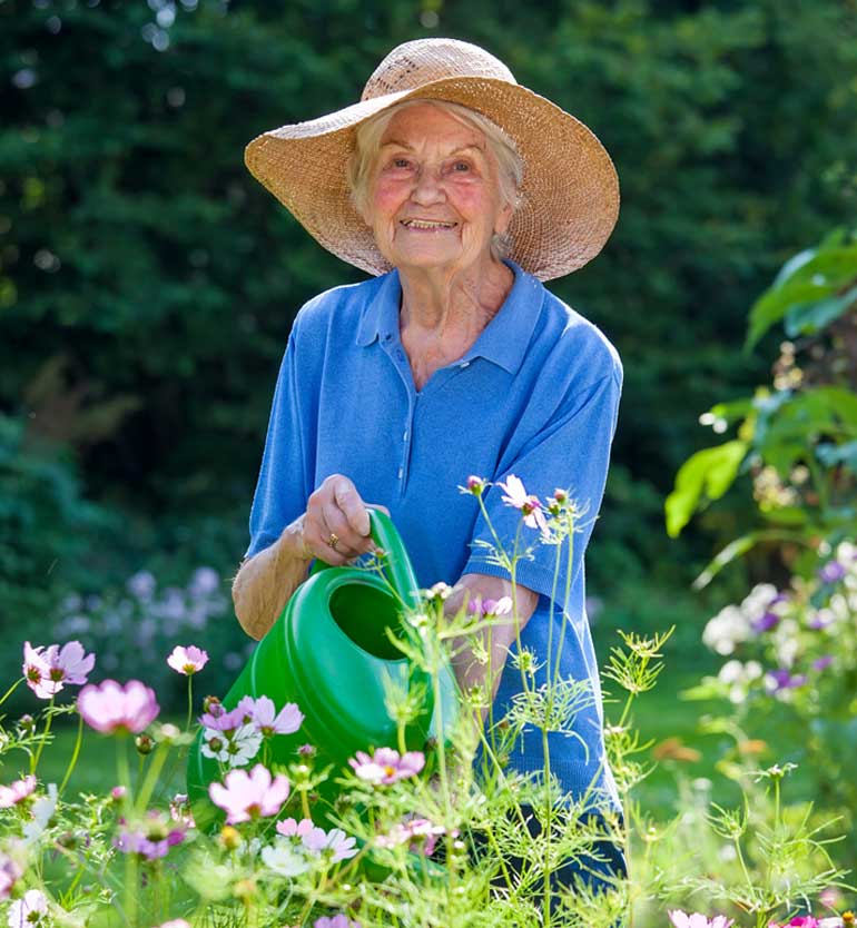 Elderly woman in straw hat watering garden flowers with a green watering can, smiling outdoors.