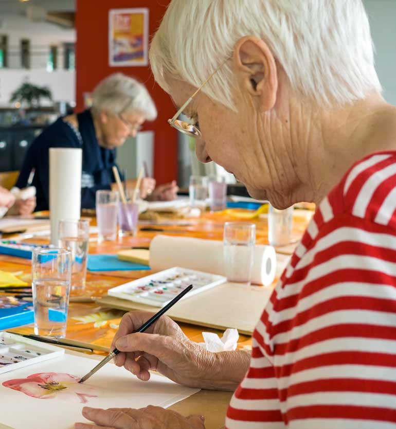 Elderly woman focusing on painting during an art class with other senior participants in the background.