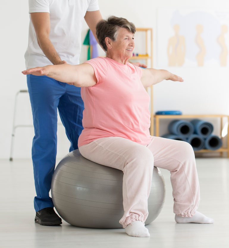 Elderly woman performs balance exercise on stability ball with therapists assistance in bright room.