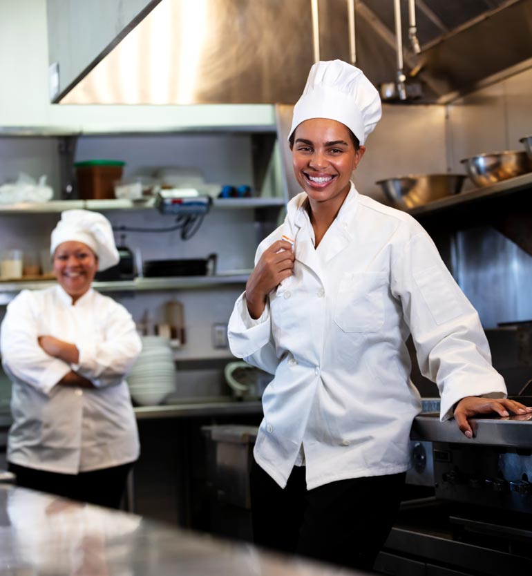 Two chefs in white uniforms and hats, smiling together in a professional kitchen.