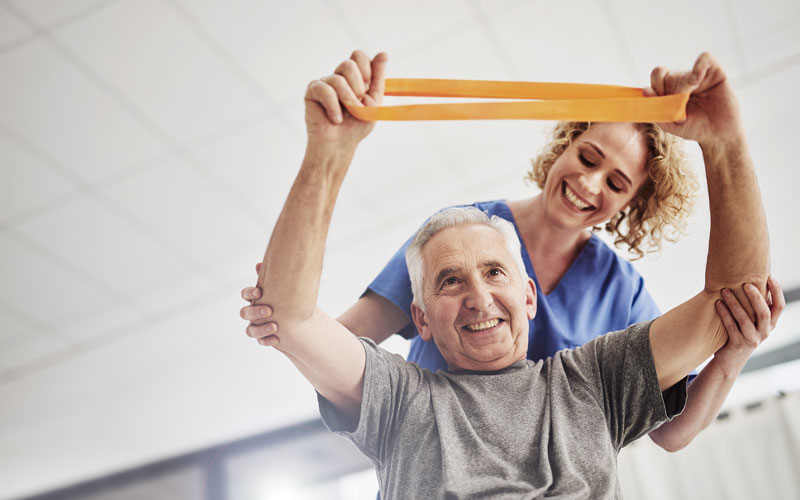 A senior man smiling and exercising with a resistance band, assisted by a supportive healthcare worker.