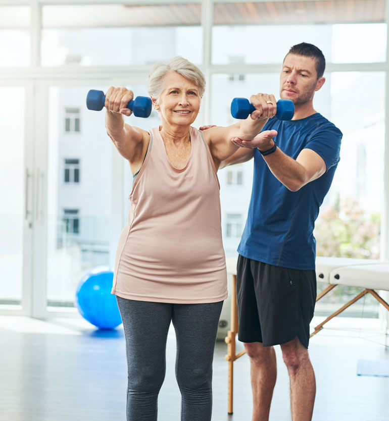 Senior woman lifting weights with the assistance of a trainer in a gym setting for fitness.